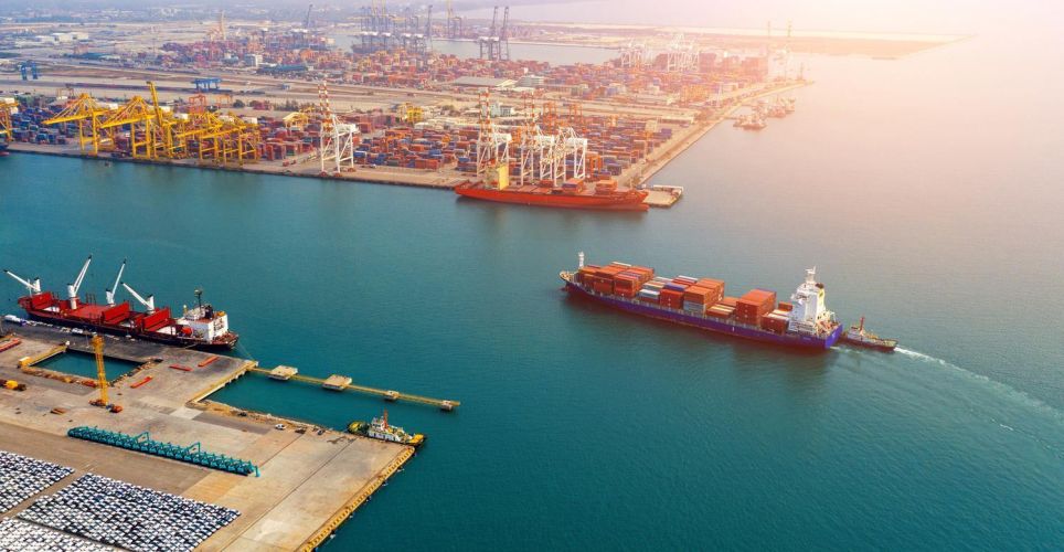 An aerial view of a port showing container ships and cranes alongside numerous containers on the quay. A container ship is leaving the harbour.