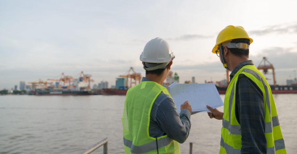 Two dockworkers look across the water at the cranes and contemplate the plan of the port terminal in their hands.