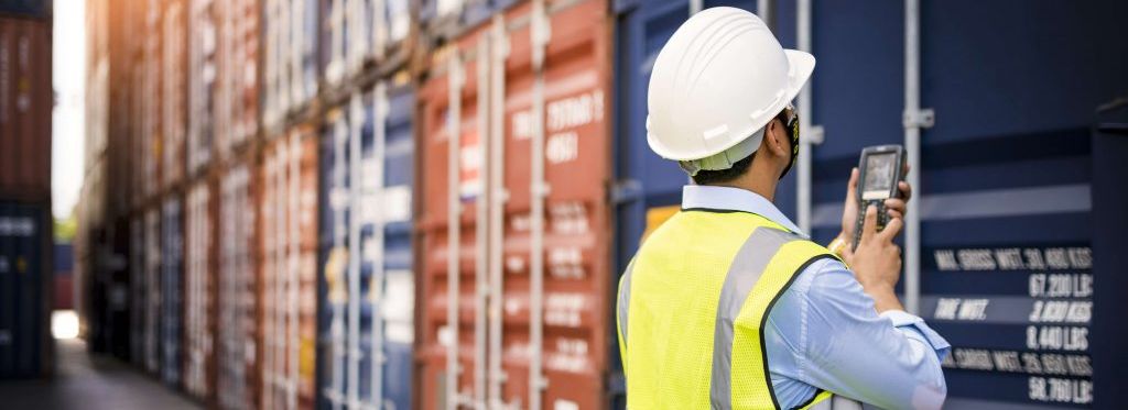 A port worker in a safety vest and white helmet stands in front of a container wall and holds a scanner in his hand.