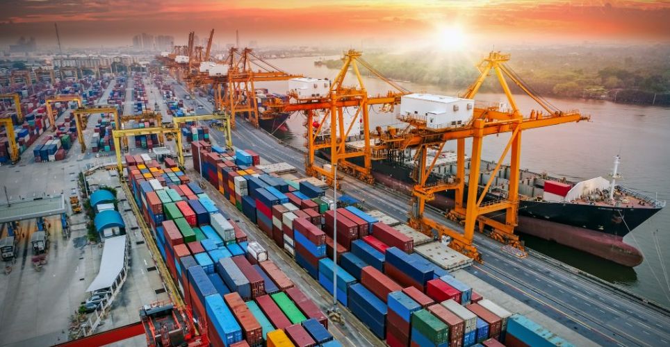 Bird's eye view of a container port with anchored ships. In the background the sun is setting.