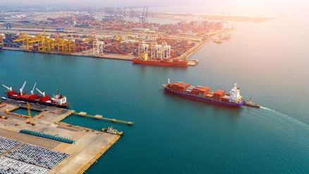An aerial view of a port showing container ships and cranes alongside numerous containers on the quay. A container ship is leaving the harbour.
