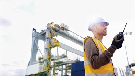 Maritime security guard with walkie-talkie in hand. Behind him part of the machines of the container terminal.